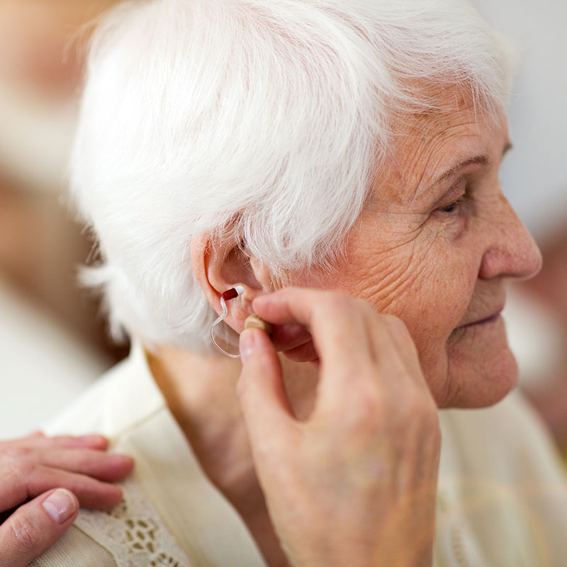 A senior man adjusts his hearing aid while a hearing aid practitioner watches.